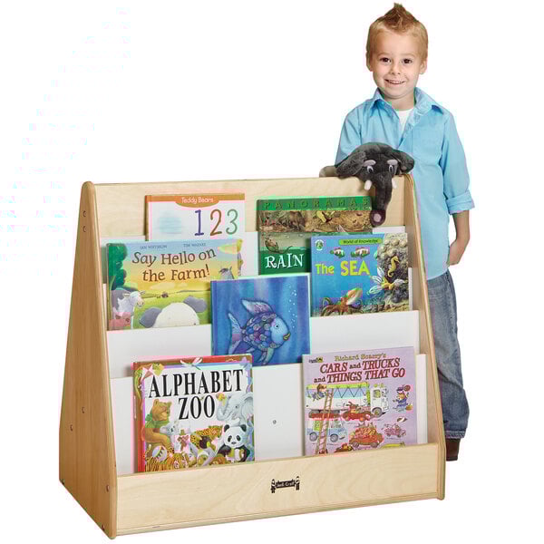A young boy standing next to a Jonti-Craft wooden book rack with many books.
