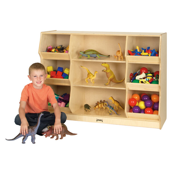 A young boy sitting next to a Jonti-Craft open storage cabinet filled with toys including a yellow dinosaur.