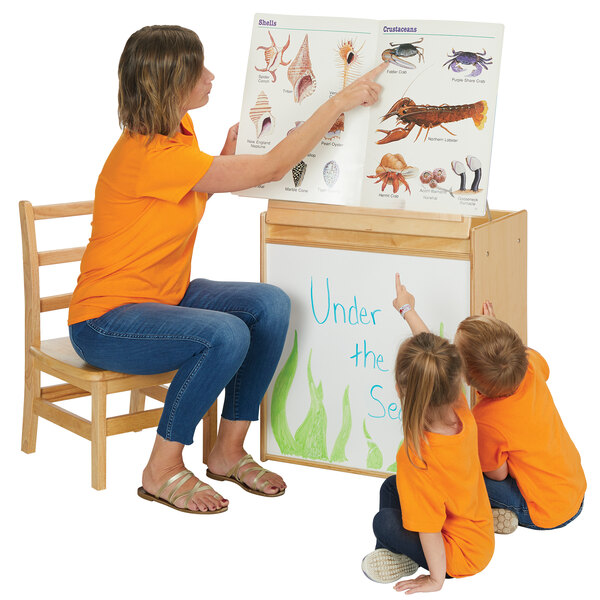 A woman sitting on a chair points at a book on a Young Time big book easel.