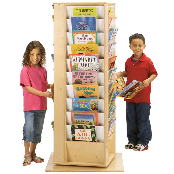 Two children standing next to a Jonti-Craft large revolving book rack filled with books.