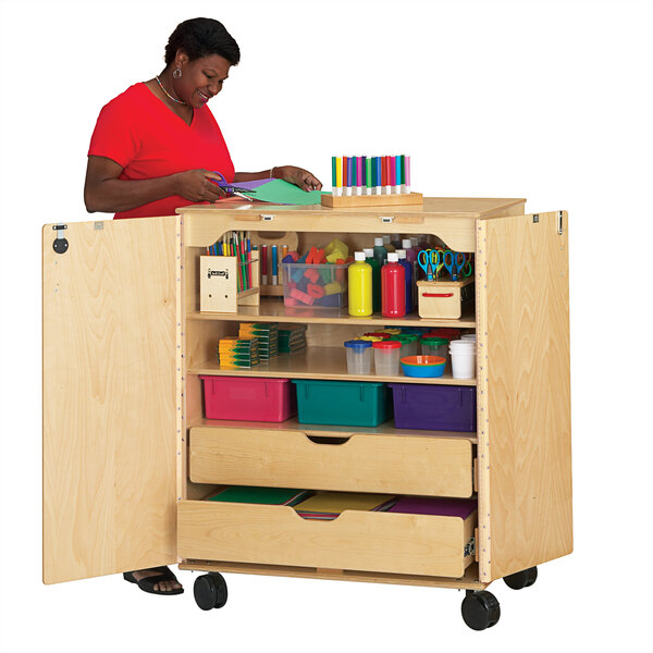 A woman standing behind a Jonti-Craft mobile wood supply cabinet filled with paper and colored objects.