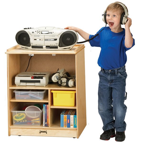 A boy wearing headphones and standing next to a Jonti-Craft wood media cart with a radio on it.