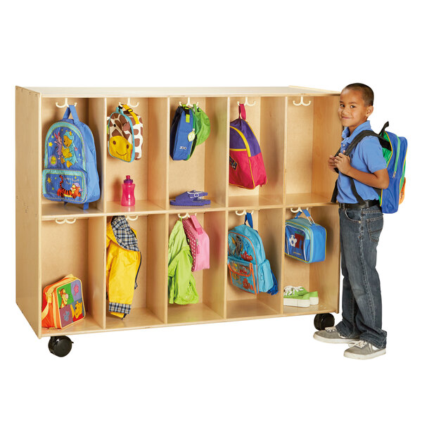 A young boy standing next to a Jonti-Craft mobile wood backpack locker with a blue backpack on a swinger.
