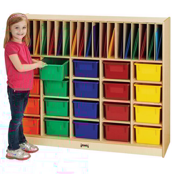 A girl standing next to a Jonti-Craft wood classroom organizer with colored cubbie trays.