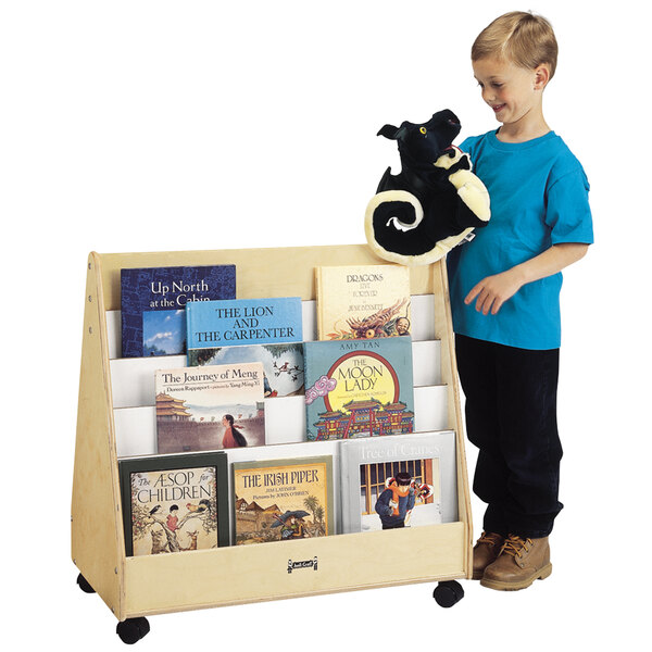 A boy standing next to a Jonti-Craft wooden double-sided book rack filled with books.