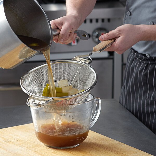 A person pouring brown liquid into a Tablecraft tin strainer over a bowl.