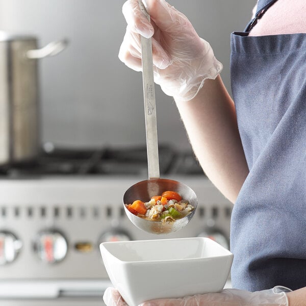 A woman in an apron using a Vollrath stainless steel ladle to serve soup over a white bowl.