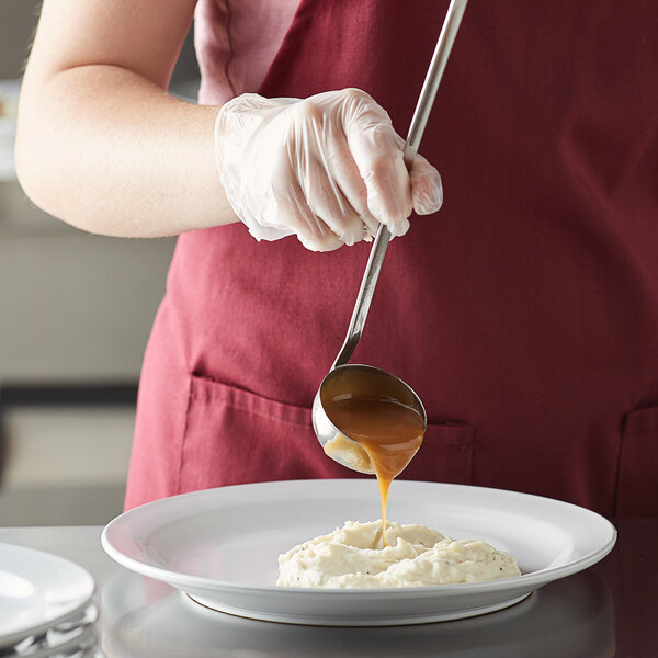 A person using a Vollrath stainless steel ladle to pour brown sauce onto mashed potatoes.