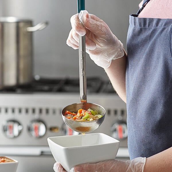 A person with a teal apron using a Vollrath stainless steel ladle to pour soup into a bowl.