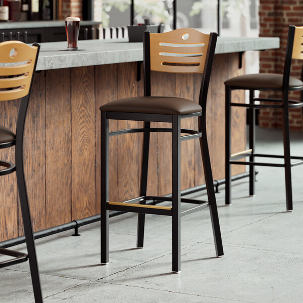 A row of Lancaster Table & Seating black restaurant bar stools with natural wood backs and dark brown vinyl seats.