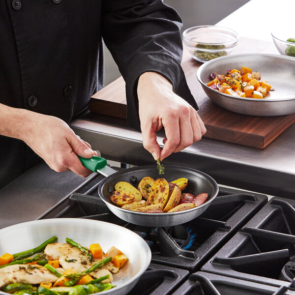 A person cooking meat, potatoes, and vegetables in a Choice aluminum non-stick fry pan with a green silicone handle on a stove.