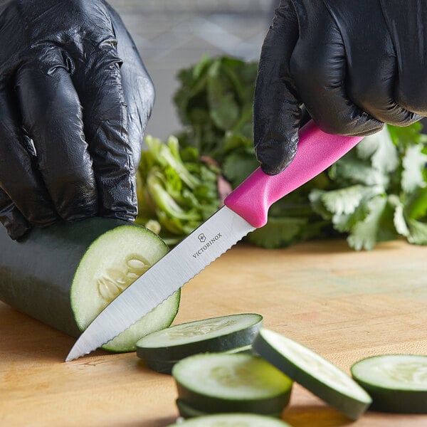 A person in black gloves using a Victorinox Swiss Classic pink paring knife to cut a cucumber on a counter.