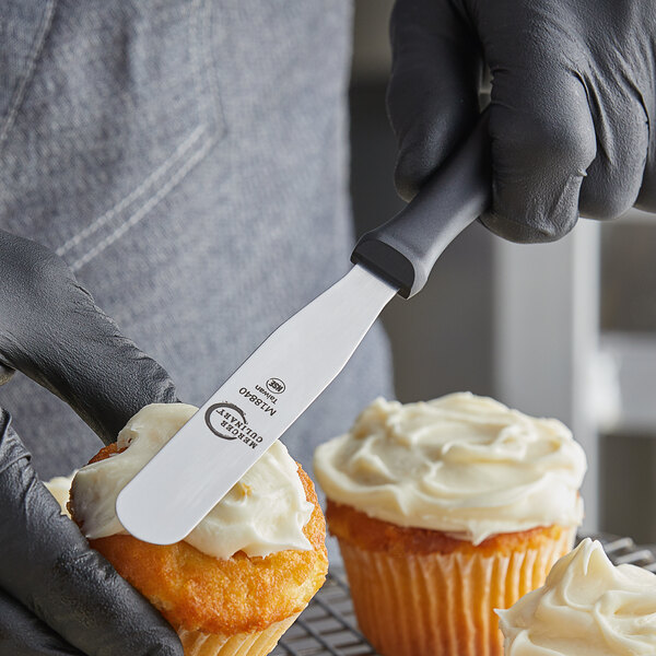 A person in black gloves using a Mercer baking spatula to frost cupcakes.