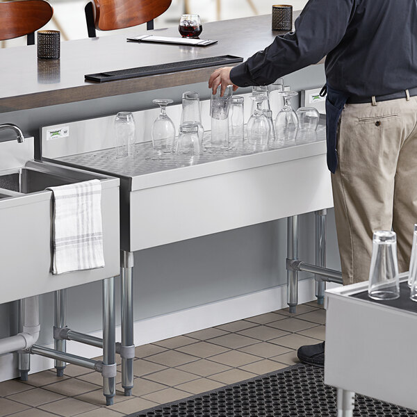 A man standing at a Regency stainless steel underbar drainboard with glasses on it.