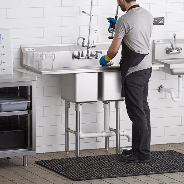 A man in a black shirt and grey pants cleaning a Regency stainless steel two compartment sink.