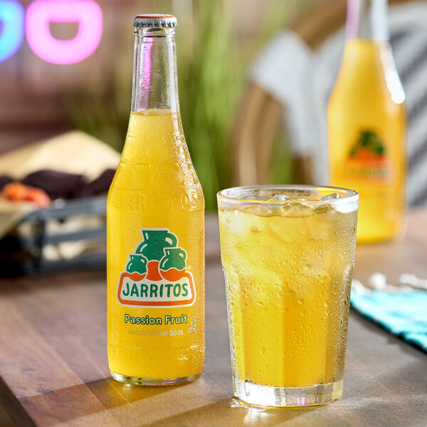A bottle of Jarritos Passion Fruit soda and a glass filled with the yellow, ice-cold beverage are placed on a wooden table.