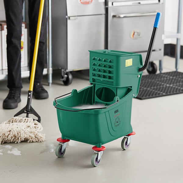 A man using a green Lavex mop bucket to clean a school kitchen floor.