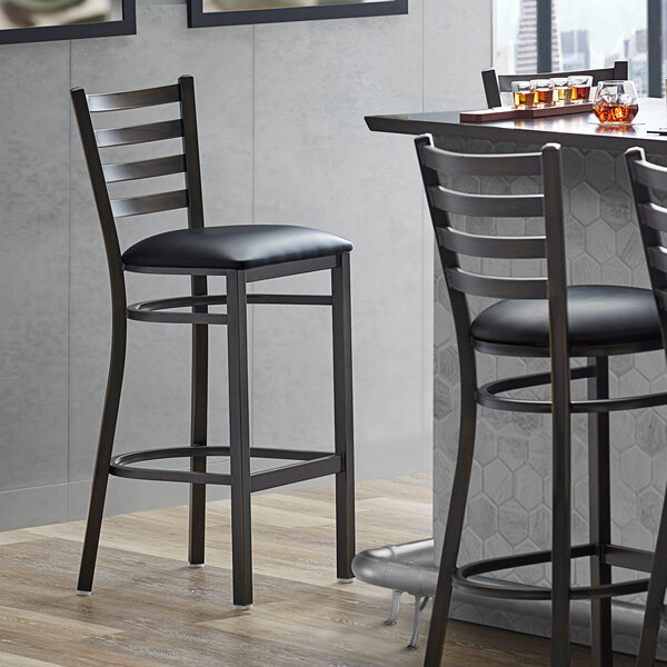 A Lancaster Table & Seating distressed copper ladder back bar stool with a black vinyl padded seat next to a table.