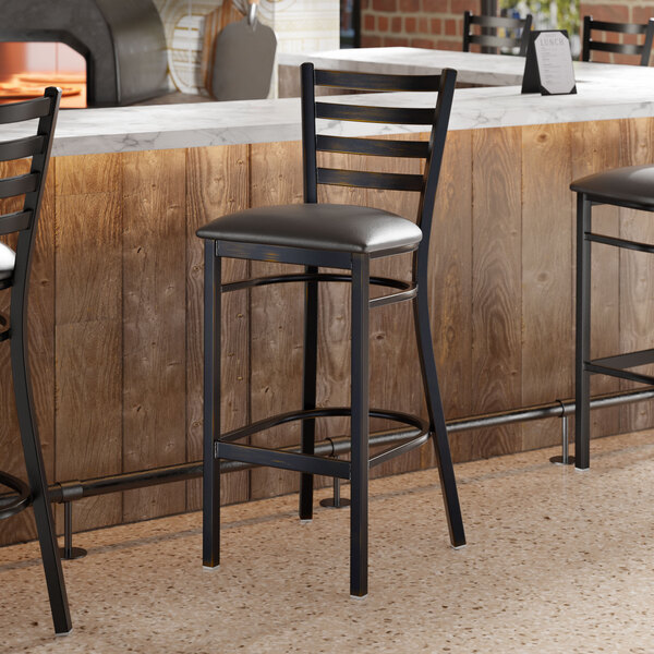 A black metal ladder back bar stool with a padded black vinyl seat and a distressed copper finish, placed at a counter.