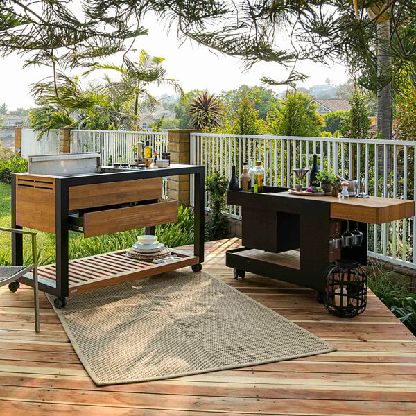 An Astella Indu+ outdoor mobile grill island with a wood and metal grill on a counter with plates and wine glasses.