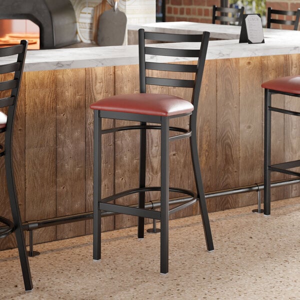 A black metal ladder back bar stool with a burgundy vinyl padded seat.