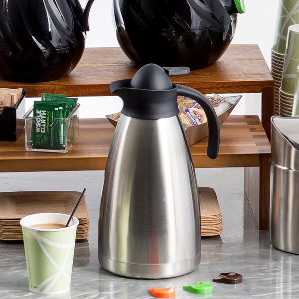 A Tablecraft stainless steel coffee carafe on a table with a cup of coffee.