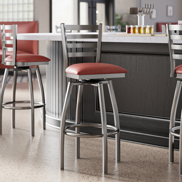A group of Lancaster Table & Seating clear coat finish ladder back swivel bar stools with burgundy vinyl padded seats next to a counter.