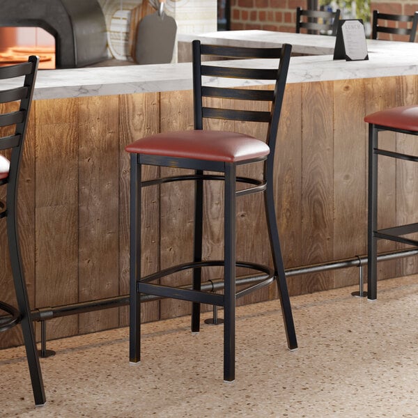 A ladder back bar stool with a distressed copper finish and a burgundy vinyl padded seat.