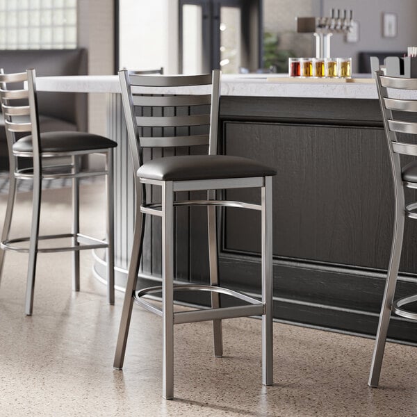 A Lancaster Table & Seating ladder back bar stool with black vinyl padded seat next to a counter.