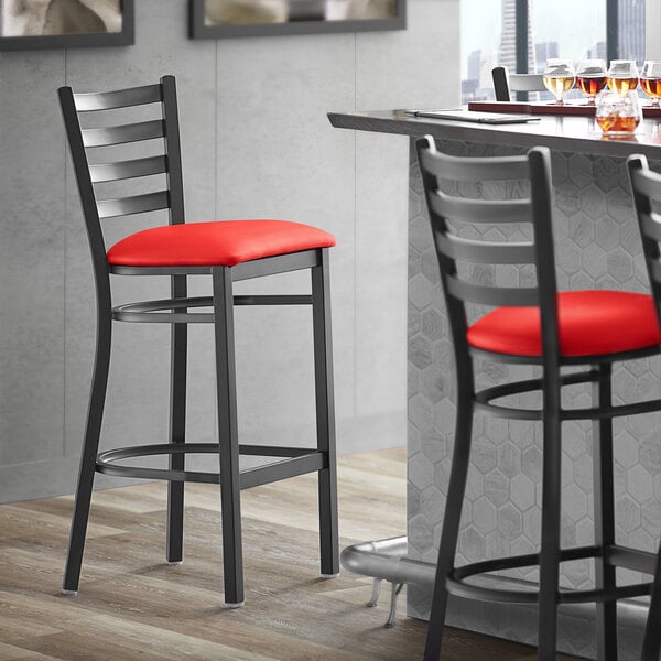 A Lancaster Table & Seating black ladder back bar stool with red vinyl padded seat.