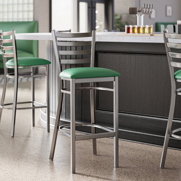 A Lancaster Table & Seating clear coat finish ladder back bar stool with a green vinyl padded seat next to a counter.