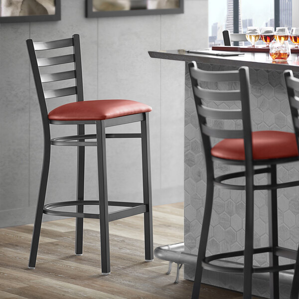 A Lancaster Table & Seating black ladder back bar stool with a burgundy vinyl cushion.