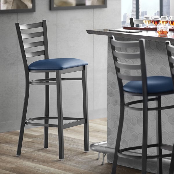 A Lancaster Table & Seating black ladder back bar stool with navy blue vinyl padded seat.