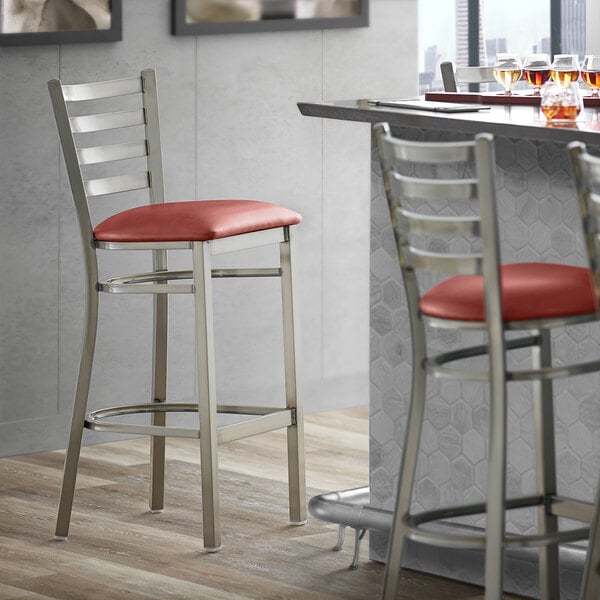 A Lancaster Table & Seating ladder back bar stool with a burgundy vinyl padded seat next to a bar counter.