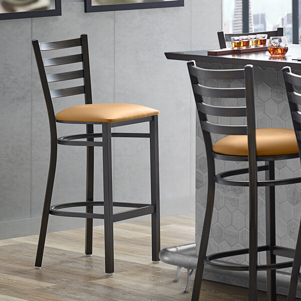 A Lancaster Table & Seating distressed copper ladder back bar stool with a light brown vinyl padded seat next to a table.