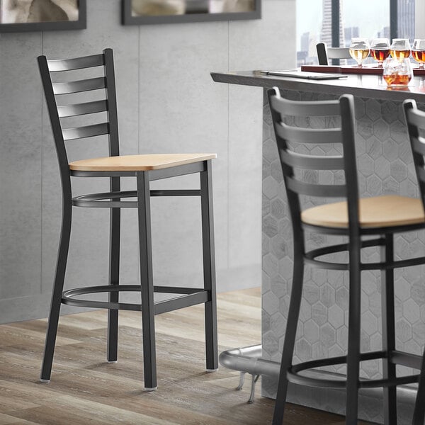 A Lancaster Table & Seating black finish ladder back bar stool with a natural wood seat next to a table.