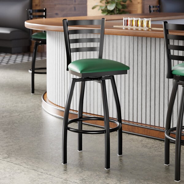 A black ladder back swivel bar stool with a green vinyl padded seat.