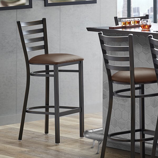 A Lancaster Table & Seating copper ladder back bar stool with dark brown vinyl cushion next to a table.