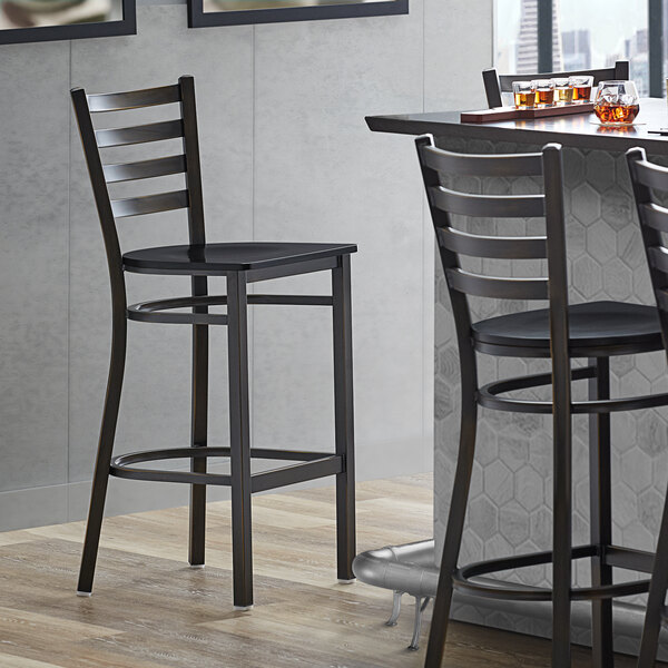 A Lancaster Table & Seating distressed copper ladder back bar stool with a black wood seat next to a table.