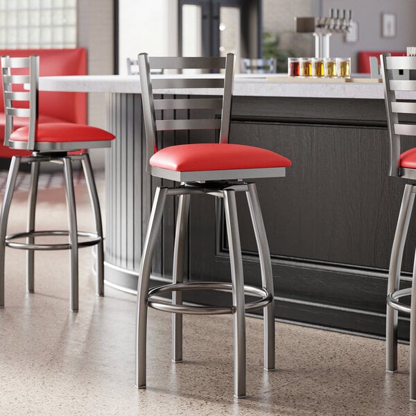 Lancaster Table & Seating Clear Coat Finish Ladder Back Swivel Bar Stool with red vinyl padded seats at a bar counter.