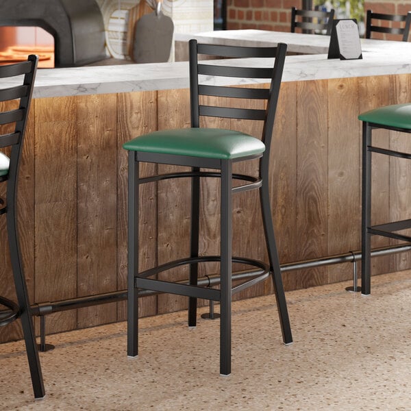 A black metal bar stool with a ladder back design and a green vinyl padded seat.
