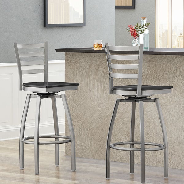 Two Lancaster Table & Seating ladder back bar stools with black wood seats and black metal legs next to a counter.