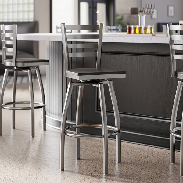 A group of Lancaster Table & Seating Clear Coat Finish Ladder Back Swivel Bar Stools with Black Wood Seats next to a counter.