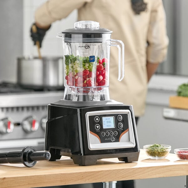A commercial blender with a digital touchpad control panel and a clear container filled with chopped vegetables, placed on a kitchen counter.