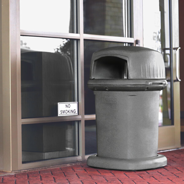 A graystone square trash can with a dome top placed outside a building entrance.