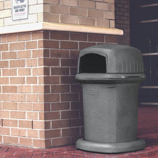 A large graystone square trash can with a dome top placed outdoors next to a brick wall.