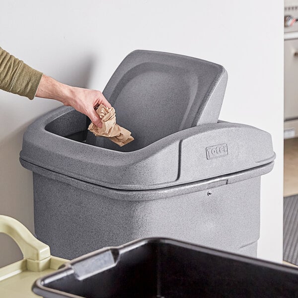A hand throwing a paper into a Toter gray square swing door lid on a trash can.