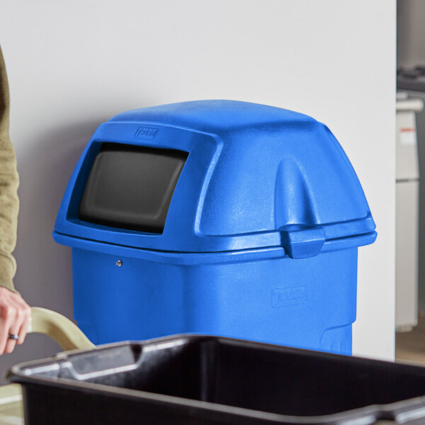 A woman standing next to a blue Toter trash can with a black swing door.