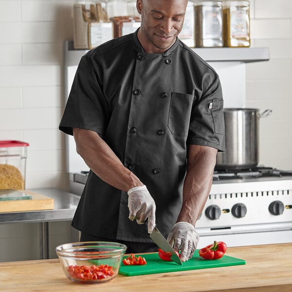 A man wearing a black Uncommon Chef short sleeve chef coat cutting vegetables.