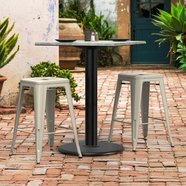 A black Lancaster Table & Seating Excalibur outdoor table base with a white stool and a black plate on a brick patio.